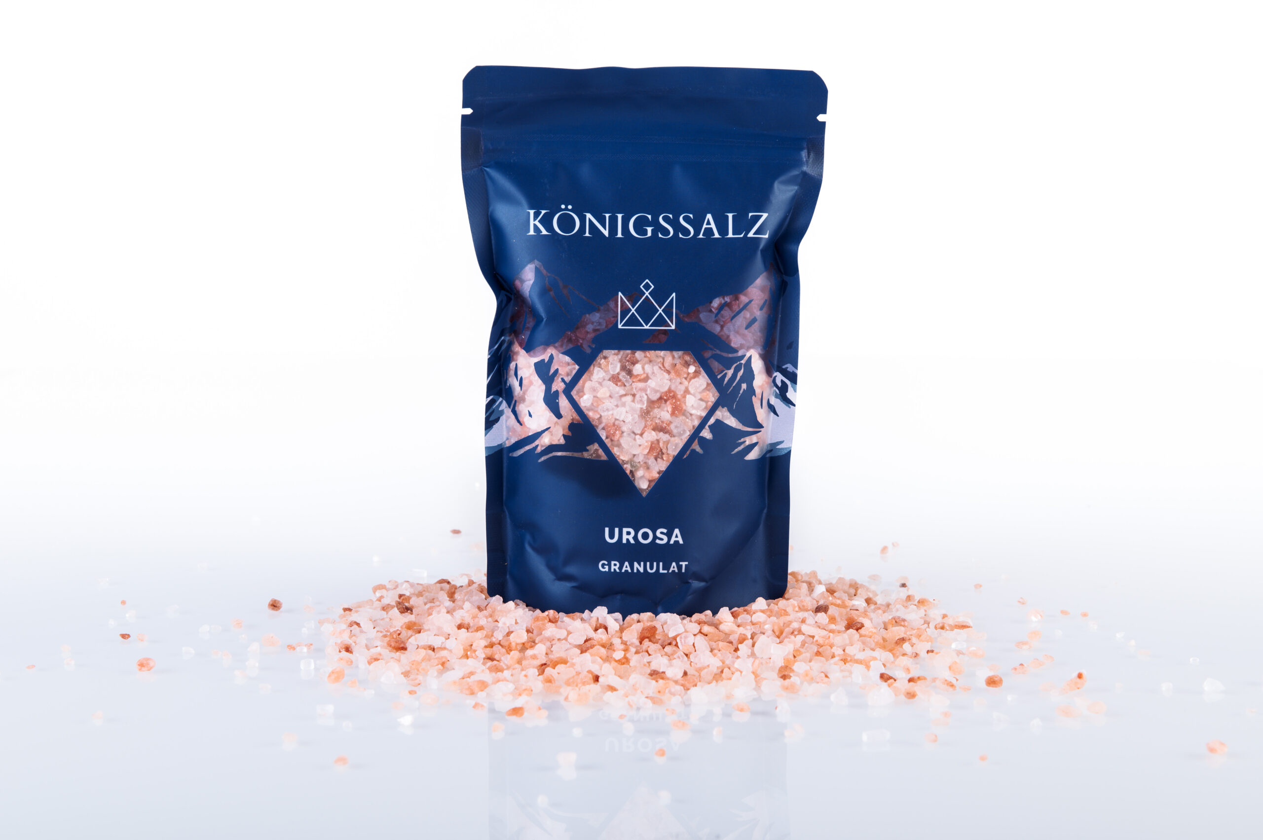 Königssalz Urosa Granulat: Rosa Himalayasalz im 500g Standbeutel auf lose verstreuten groben Salzkristallen vor weißem Hintergrund